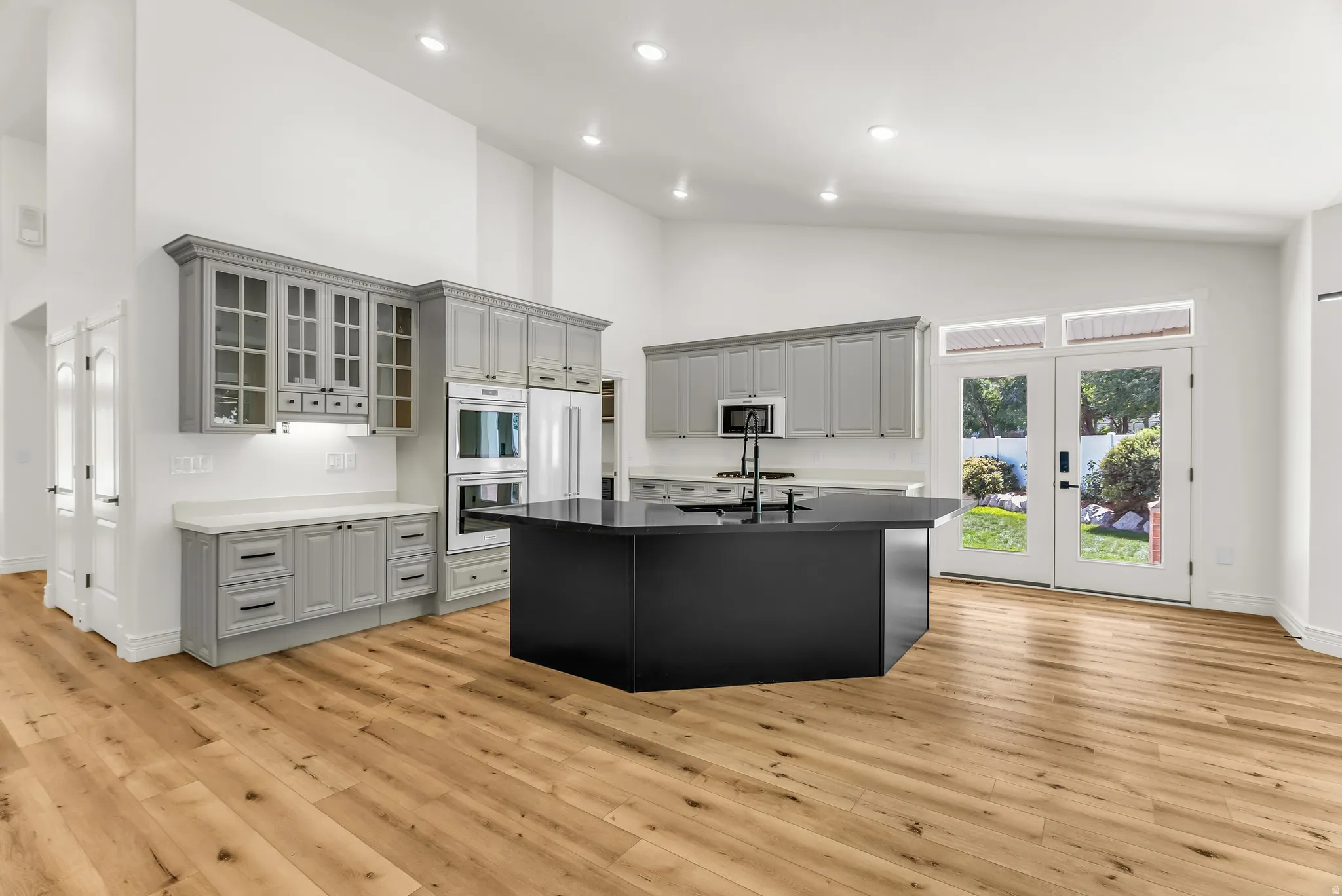 Vaulted ceilings with recessed lighting, light wood-finish flooring, custom cabinetry with glass-front uppers, built-in double oven, and double French patio doors