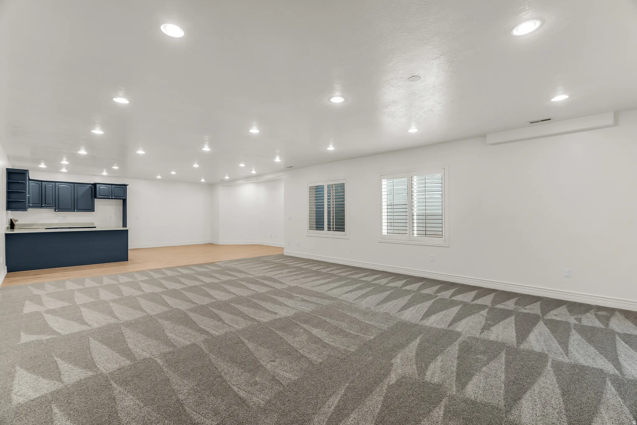 Spacious room featuring recessed lighting, a built-in wet bar with dark cabinetry and a light-toned countertop, two windows with louvered blinds, and a combination of patterned carpeting and wood-finish flooring
