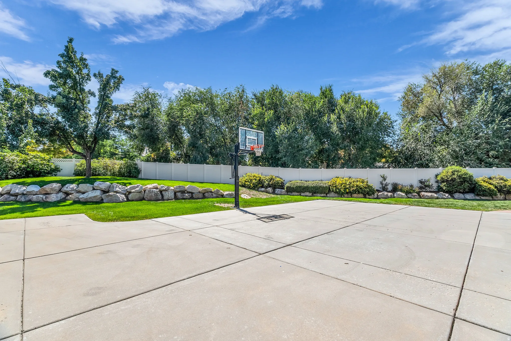 Expansive concrete patio with a basketball hoop, surrounded by a manicured lawn, landscaping, and a privacy fence
