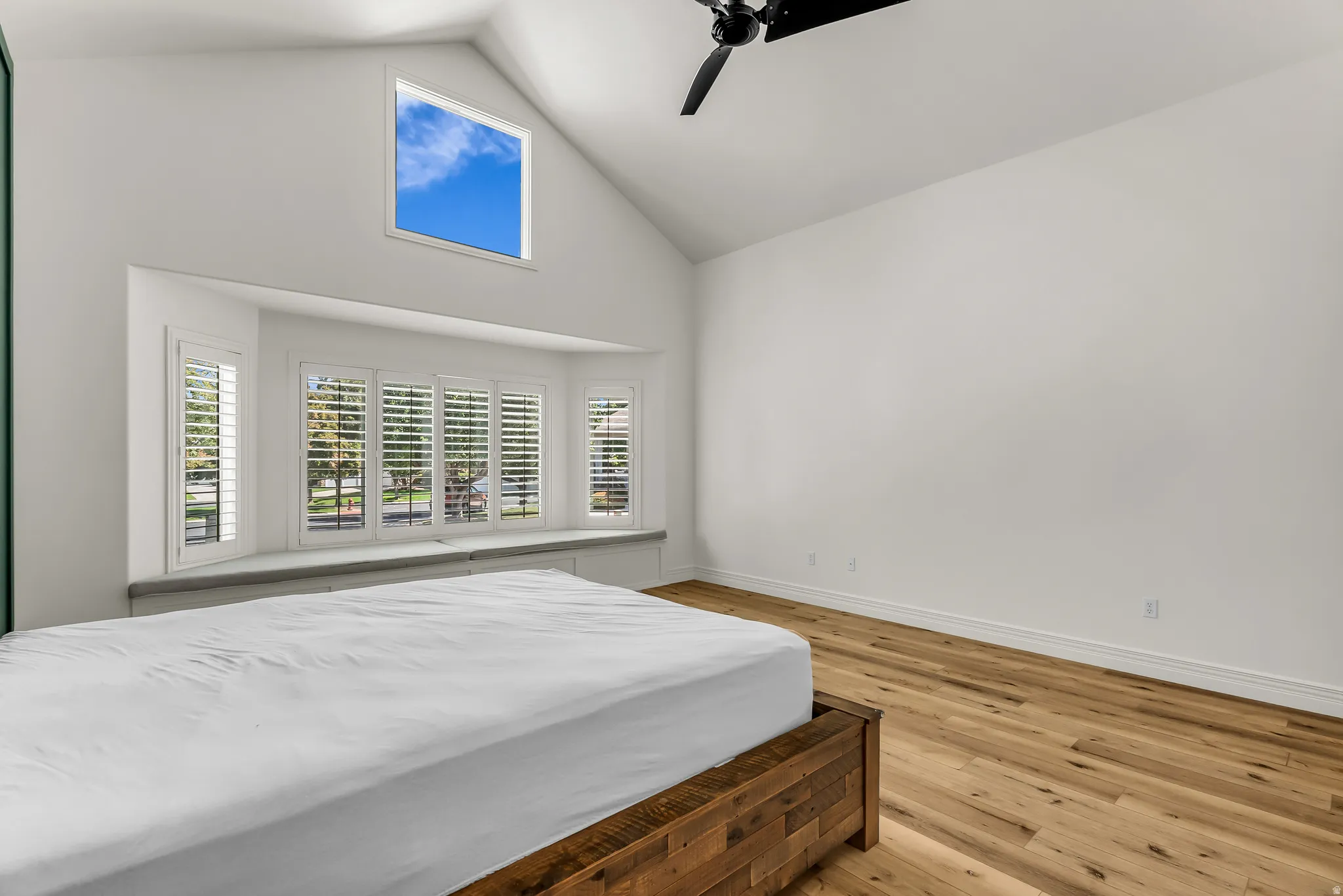 Vaulted ceiling with a ceiling fan, wood-finish flooring, a bay window with plantation shutters, and an upper transom window