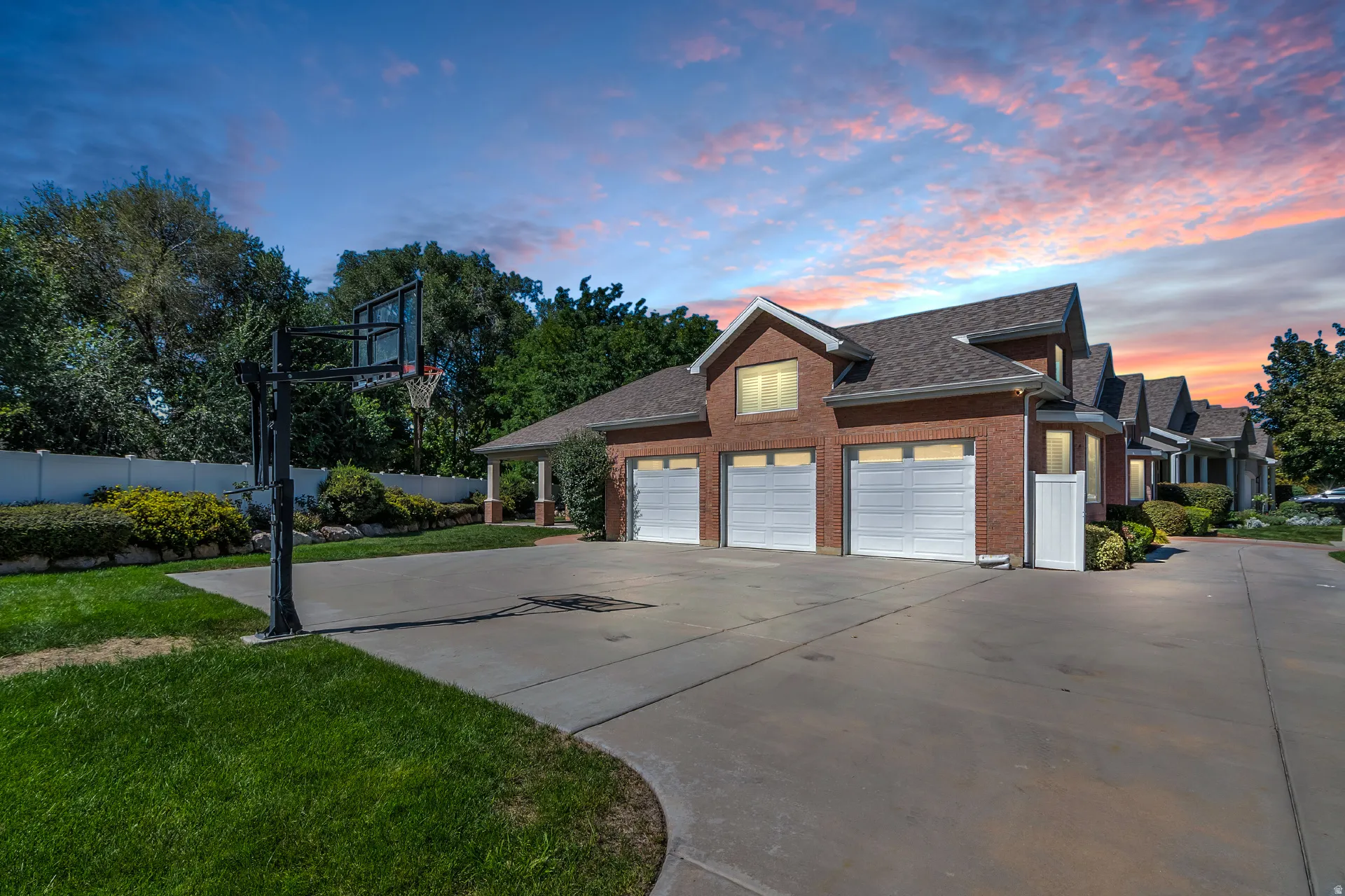 Brick exterior with three garage doors, a concrete driveway, a basketball hoop, and established landscaping