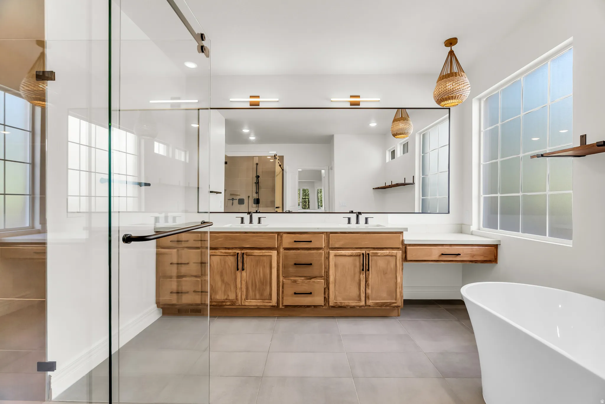 Dual wood-finish vanities with countertop, expansive vanity mirror, walk-in shower with clear glass enclosure, freestanding soaking tub, and large grid-pattern windows