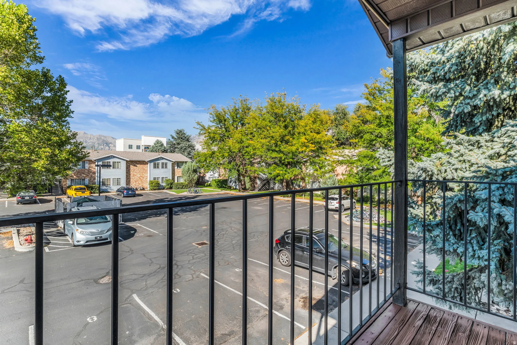 Balcony with wood-finish decking and black metal railing overlooking a parking area with mature trees and distant mountains