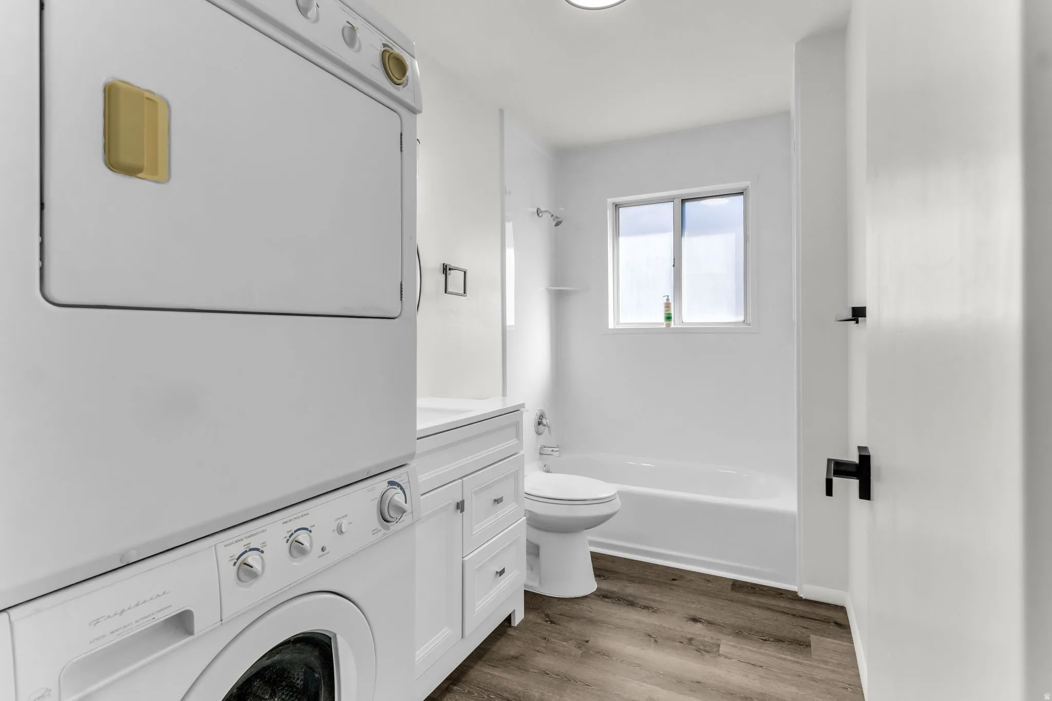 Integrated laundry appliances, white cabinet vanity, white bathtub with shower, white toilet, and wood-finish flooring