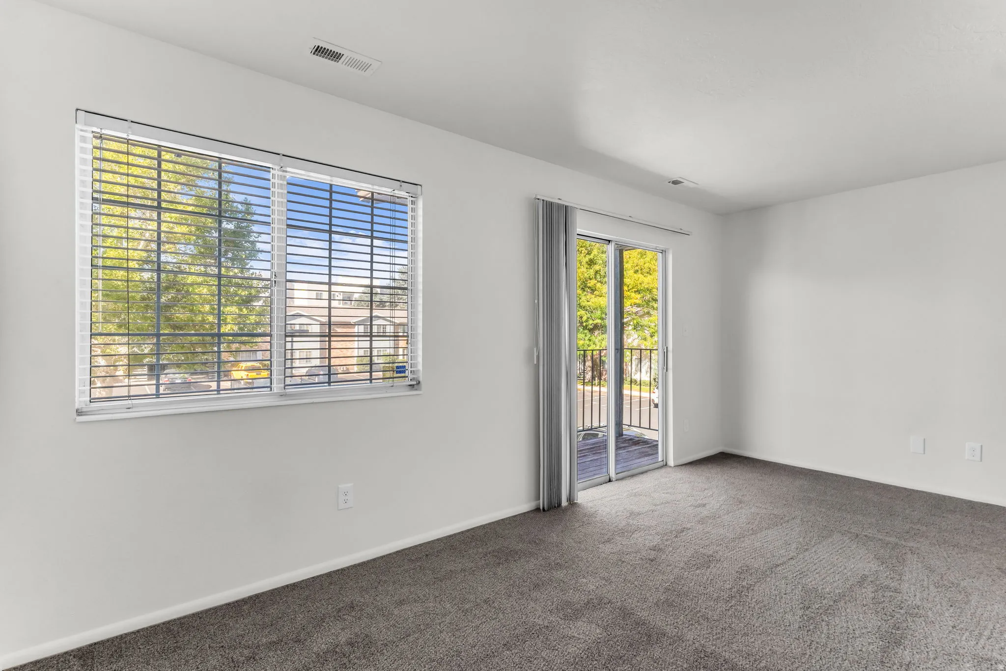Expansive room featuring light gray carpeting, crisp white walls, a large window with blinds, and a sliding glass door leading to an outdoor space