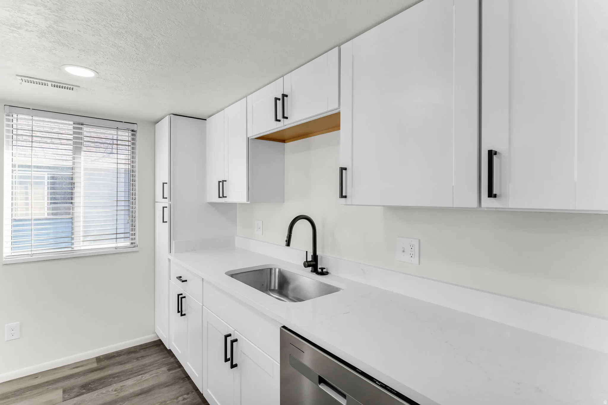 White shaker-style cabinetry with matte black hardware, stainless steel sink with a matte black faucet, integrated dishwasher, and wood-finish flooring