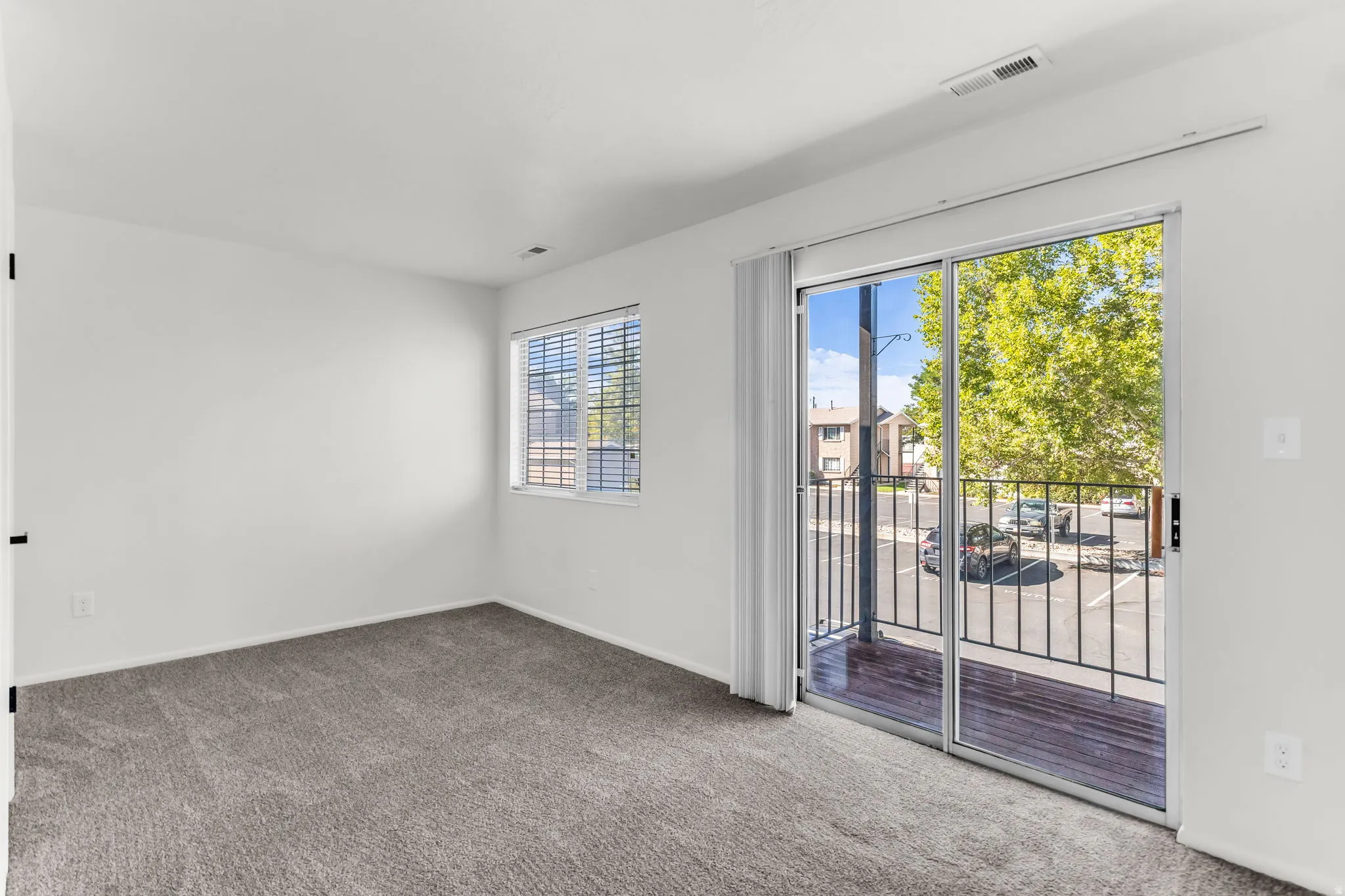 Sliding glass door leading to a balcony with metal railings, a window with blinds, and gray carpet