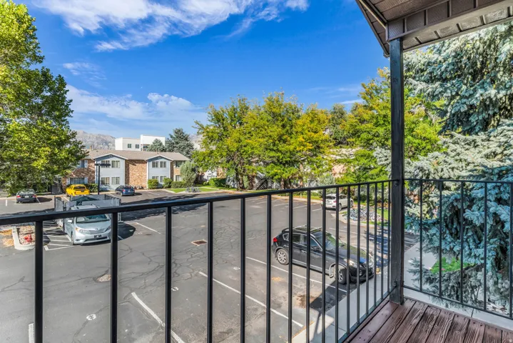 Balcony with wood-finish decking and black metal railing overlooking a parking area with mature trees and distant mountains