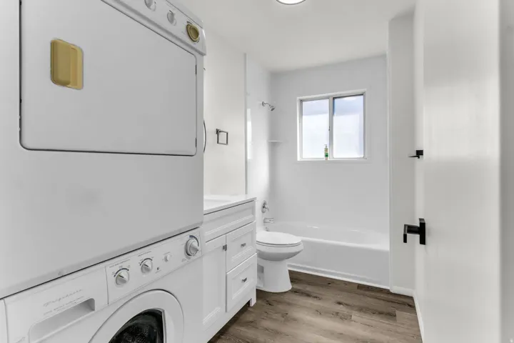 Integrated laundry appliances, white cabinet vanity, white bathtub with shower, white toilet, and wood-finish flooring