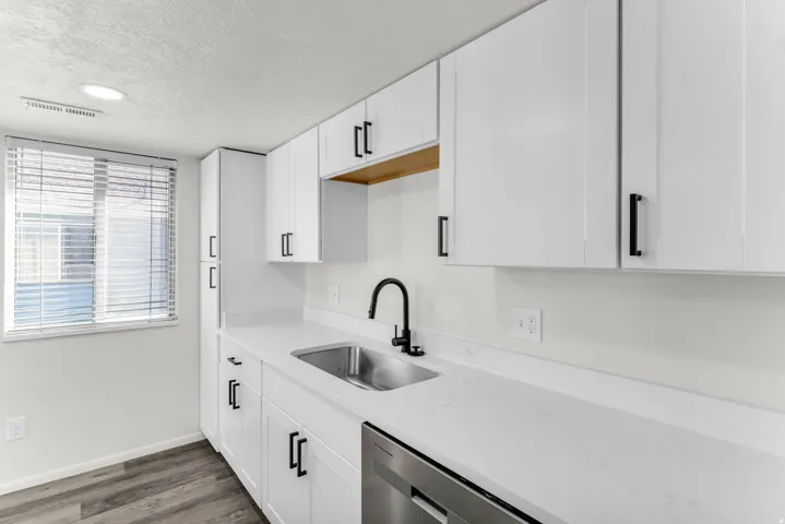 White shaker-style cabinetry with matte black hardware, stainless steel sink with a matte black faucet, integrated dishwasher, and wood-finish flooring