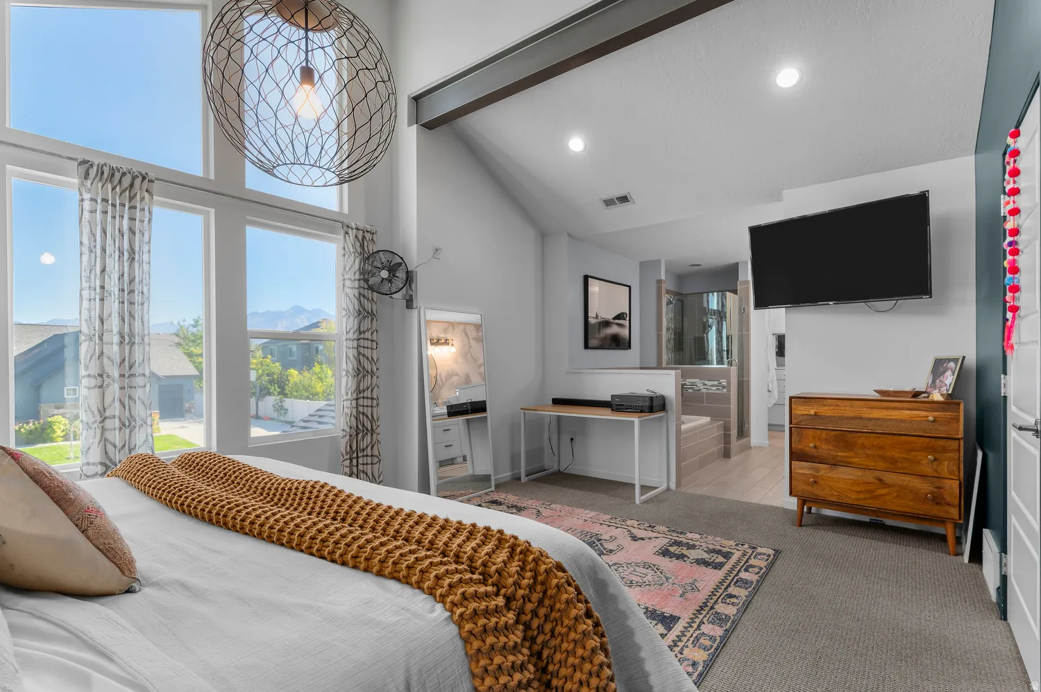 Vaulted ceiling with exposed beam, expansive window array, contemporary pendant light fixture, recessed lighting, and an open-concept bathroom area with a built-in tub