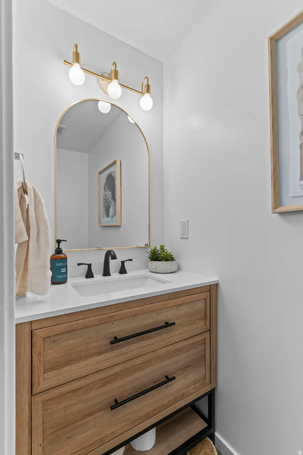 Wall-mounted three-light fixture with a gold-tone finish, arched mirror, single-basin vanity with a white countertop, wood-finish cabinetry, and matte black faucet