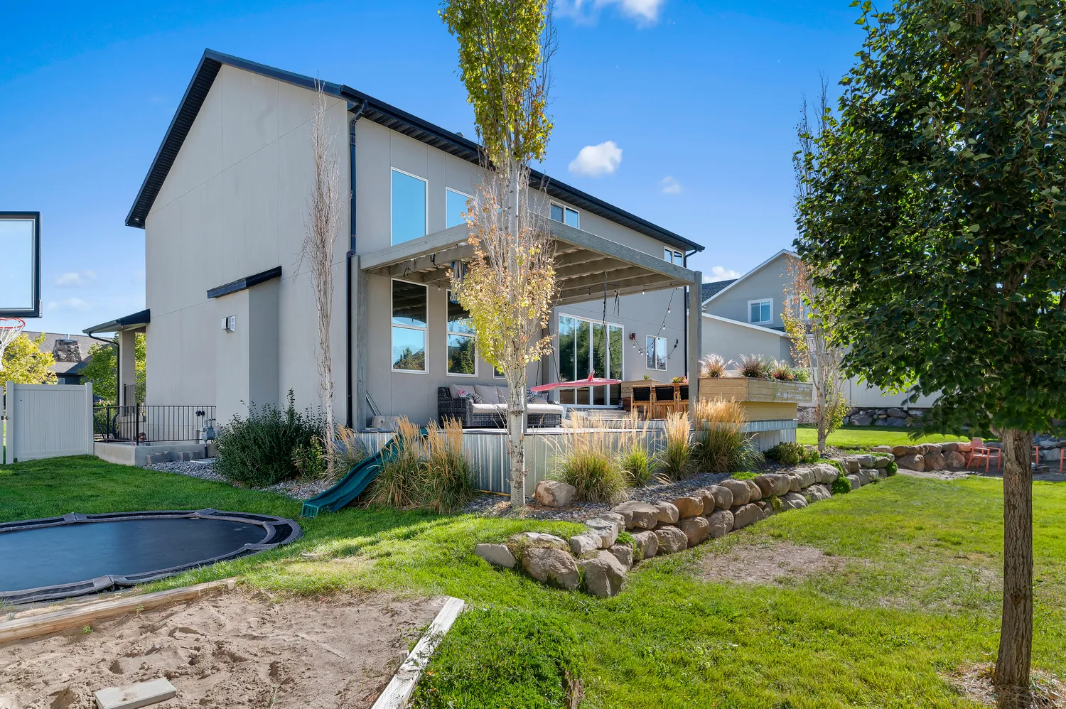 Expansive backyard featuring a manicured lawn, established landscaping, natural stone retaining walls, and a large elevated patio with a pergola