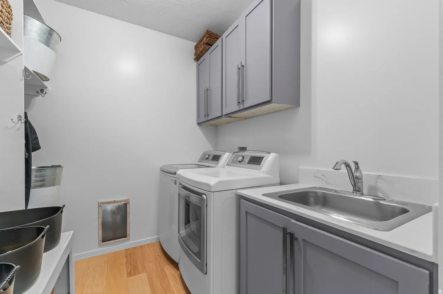 Light wood-finish flooring, gray upper and lower cabinetry, stainless steel sink with chrome faucet, and white countertop