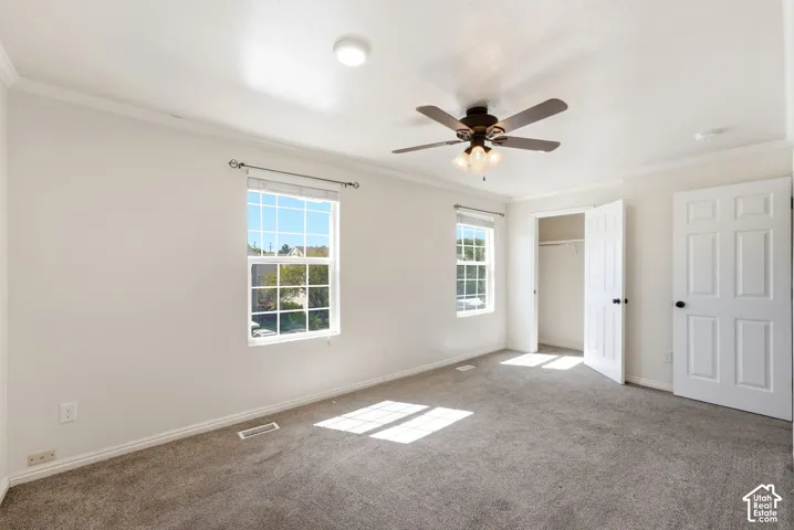 Neutral tone carpeted flooring, two windows with blinds, a ceiling fan with integrated lighting, a flush-mount ceiling light, and crown molding
