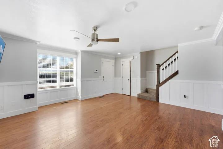 Wood-finish flooring, white panel wainscoting, a ceiling fan with light, crown molding, and a staircase with wood newel post and handrail
