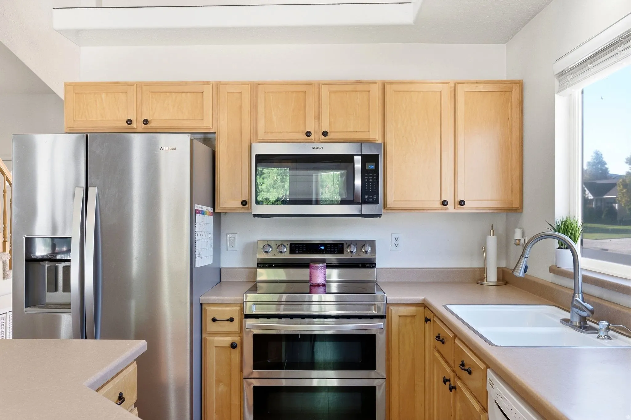 Stainless steel side-by-side refrigerator, stainless steel double oven range, stainless steel microwave, light wood-finish cabinetry, and a single basin sink