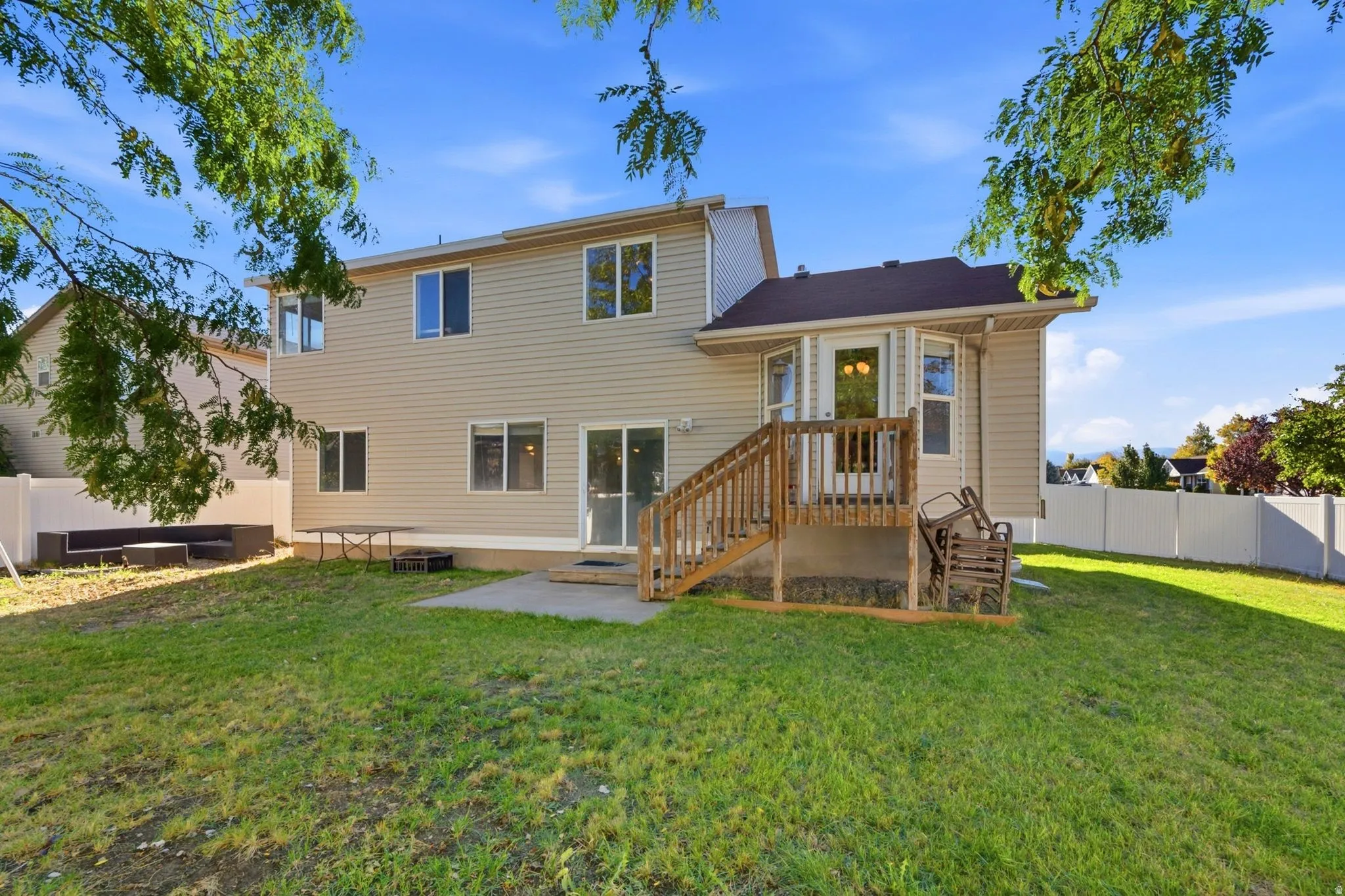 Expansive grassy yard with a concrete patio, wood deck with stairs, multiple windows, and a sliding glass door