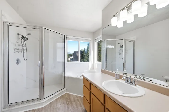 Walk-in shower with clear glass enclosure, built-in bathtub, dual-sink vanity with light-colored countertop, wood-finish cabinetry, and wood-finish flooring