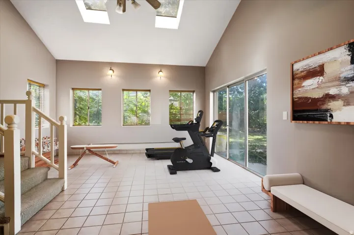 Sliding glass door, multiple windows with blinds, tiled flooring, vaulted ceiling with skylights, and a wood staircase with carpeted treads