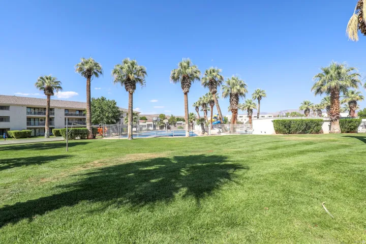 Expansive green lawn with mature palm trees, multi-story buildings with balconies, and a fenced recreational court