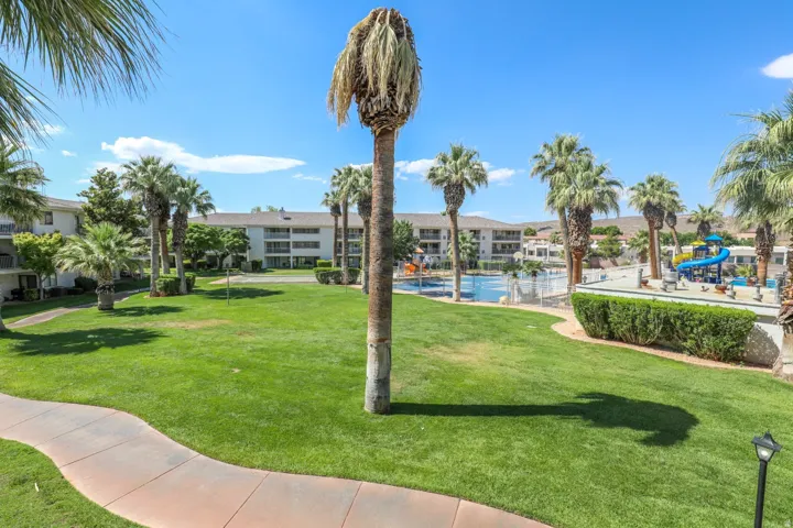 Expansive green space featuring a paved pathway, numerous palm trees, distant multi-story buildings, and a clear blue sky