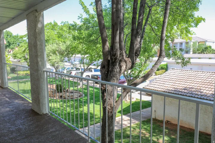 Covered outdoor walkway with white metal railings, overlooking a manicured lawn, established trees, and stucco buildings with tile roofs