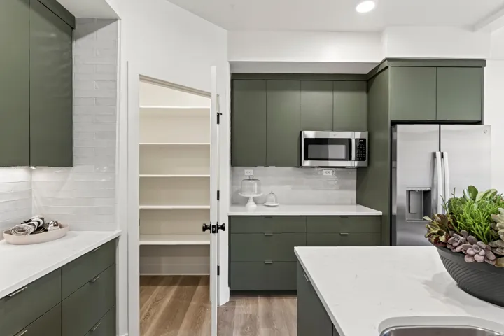 Modern kitchen featuring green flat-panel cabinetry, white countertops, stainless steel appliances, a white subway tile backsplash, and a walk-in pantry with wood-finish flooring
