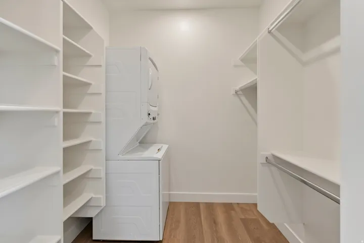 White shelving units, built-in clothing rods, stacked washer and dryer appliances, and wood-finish flooring