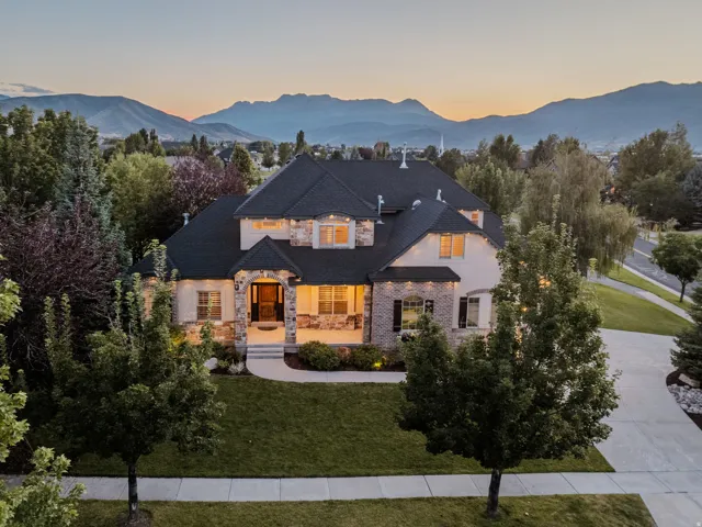Stucco exterior with stone accents, dark shingles, an inviting covered porch, and mature landscaping