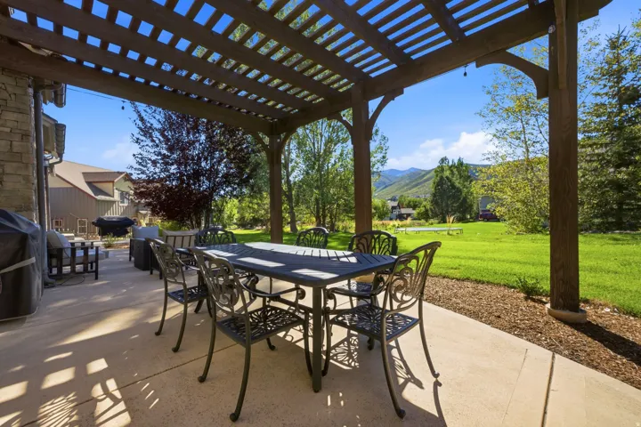 Expansive concrete patio featuring a wood pergola, stone exterior wall, and a large grassy area with mature trees