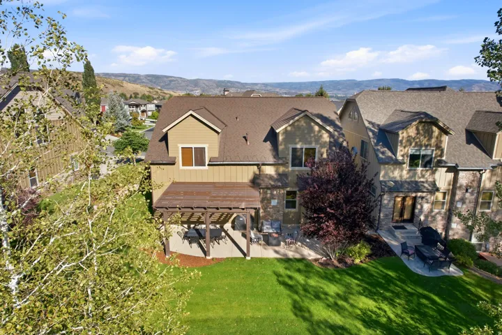 Expansive green lawn, stone patio, attached pergola, mature landscaping, and multi-story architectural design