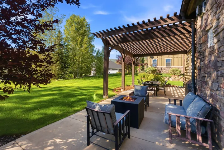 Expansive concrete patio with a wooden pergola, stone-clad exterior, and a large grassy yard with mature trees