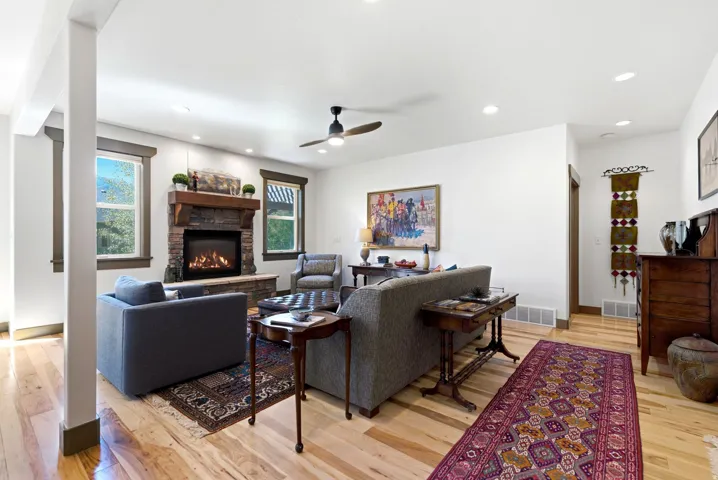 Hardwood flooring throughout, stone-clad fireplace with wood mantle, recessed lighting, ceiling fan, and large windows with natural light