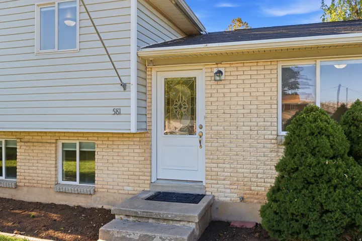 Split-level exterior featuring light-colored brick and horizontal siding, a white front entry door with decorative glass, white-framed windows, and mature foundation plantings