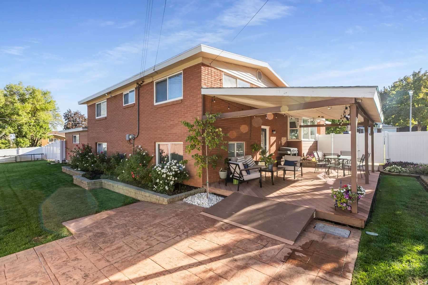 Back of the home.Red brick exterior with white trim, covered wood deck, stamped concrete patio, and extensive landscaping with flowering shrubs