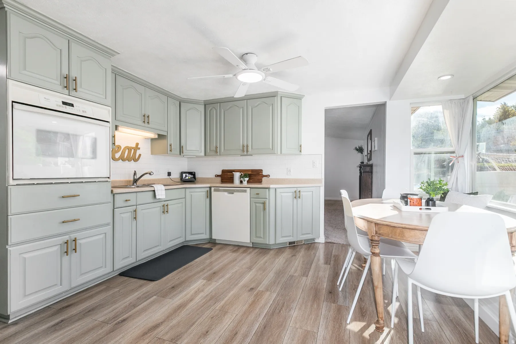 Built-in wall oven, light-toned cabinetry with gold-toned hardware, wood-finish flooring, white subway tile backsplash, and a ceiling fan