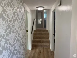 Hallway with wood-finish flooring, patterned accent wall, and carpeted stairs with a dark handrail