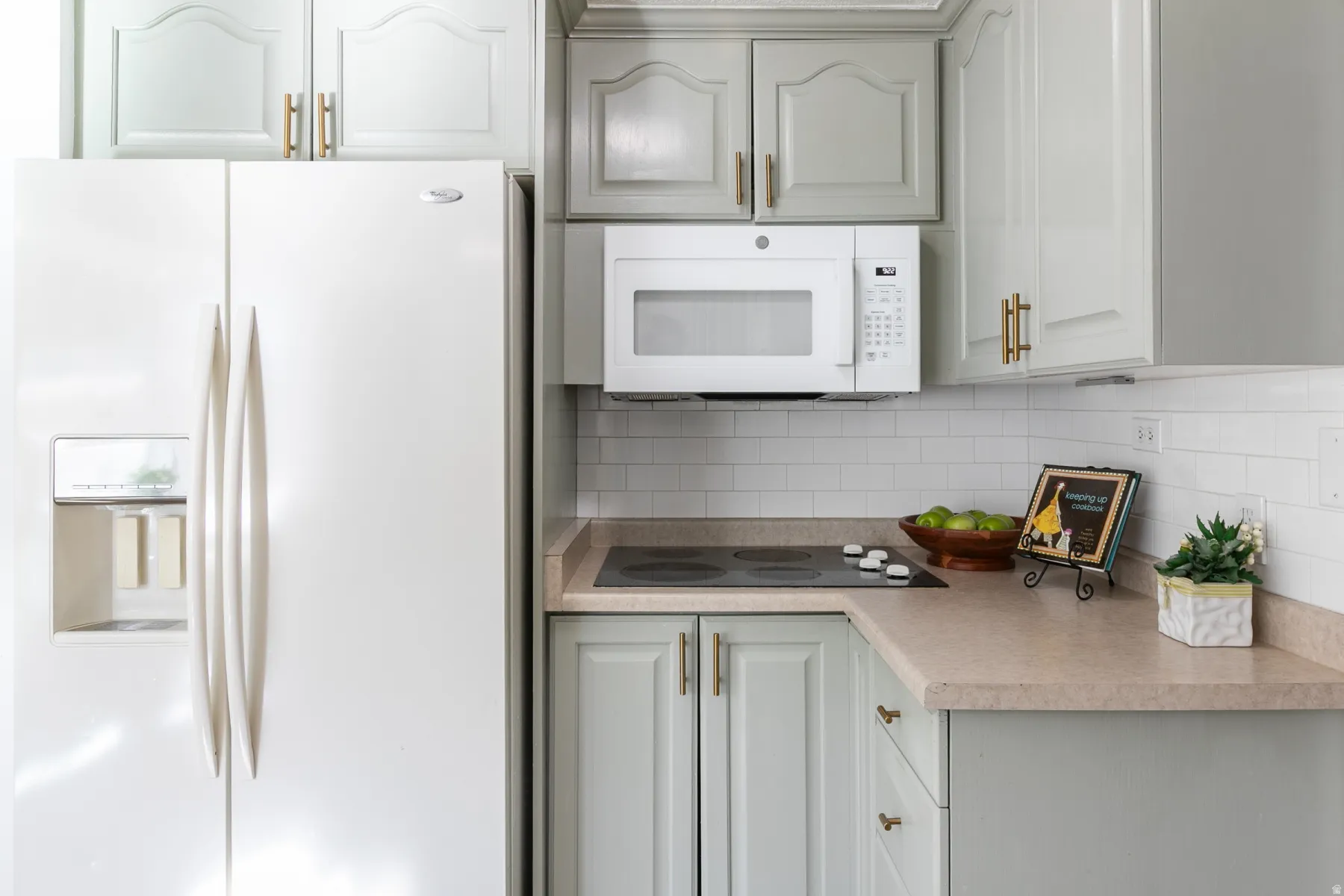 White subway tile backsplash, light-toned countertops, white built-in microwave, light-colored cabinetry with gold-toned hardware, and a white side-by-side refrigerator with an external dispenser