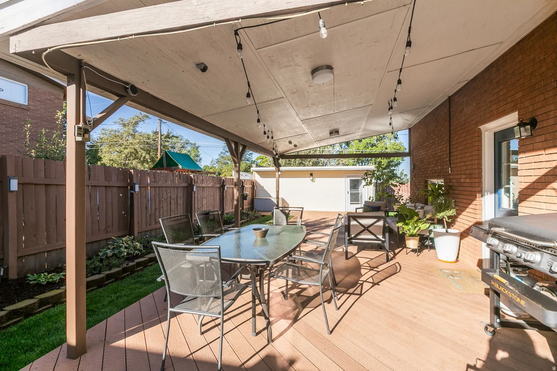 Covered outdoor deck with string lighting, brick exterior wall,  wood privacy fence and flower garden.