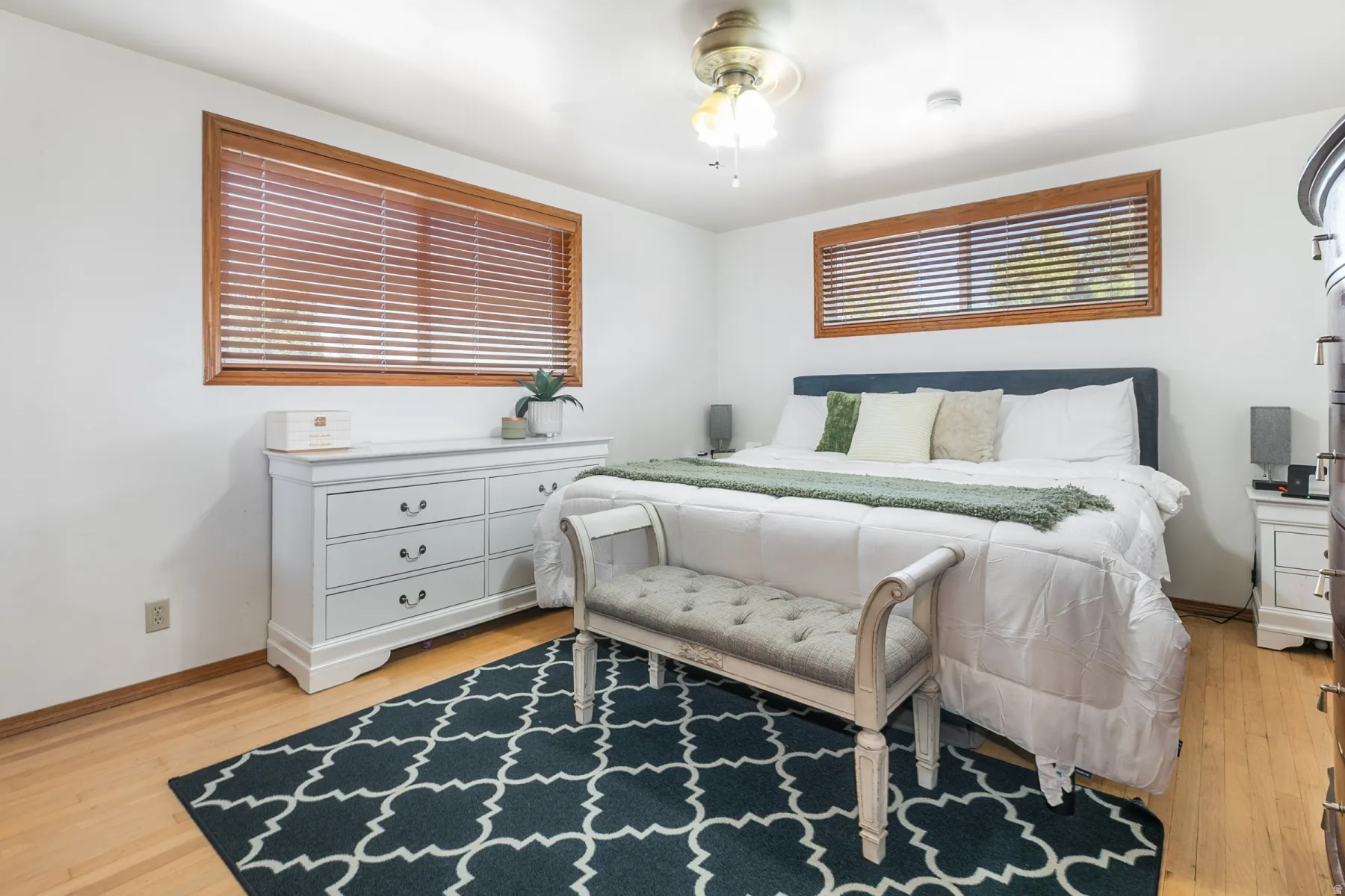 Wood-finish flooring, white walls, two windows with wood trim and horizontal blinds, and a ceiling fan with integrated lighting