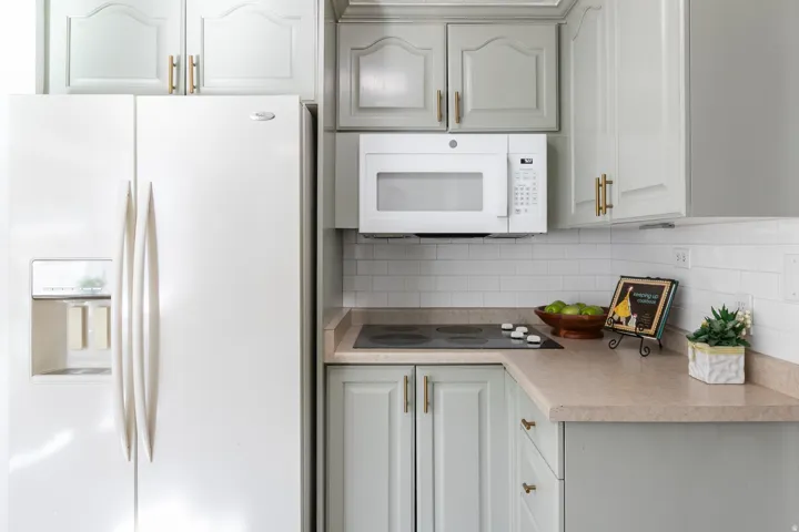 White subway tile backsplash, light-toned countertops, white built-in microwave, light-colored cabinetry with gold-toned hardware, and a white side-by-side refrigerator with an external dispenser
