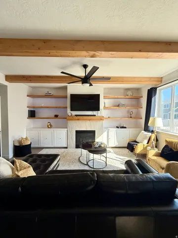 Exposed wood ceiling beams, built-in shelving with integrated lighting, fireplace with stone surround, built-in cabinetry, and a ceiling fan with integrated lighting