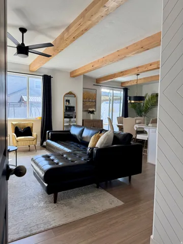 Wood-finish flooring, exposed ceiling beams, modern ceiling fan, large windows with dark drapery, and a decorative accent wall