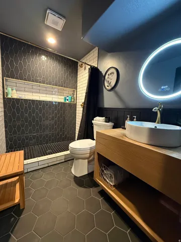 Black hexagonal floor tiling, wood-finish vanity with an integrated basin sink, black vertical shiplap paneling, black hexagonal shower wall tiling, and a recessed shower niche