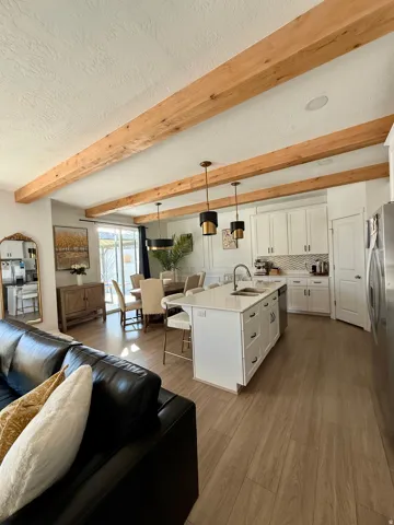 Wood-finish flooring throughout, kitchen island with built-in sink and seating, white cabinetry with dark hardware, recessed lighting, and exposed wooden ceiling beams