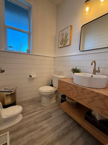 Wood-finish flooring, shiplap wall paneling, modern vanity with open shelving, vessel sink, and wall-mounted light fixture