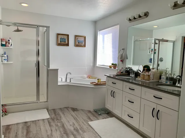 Wood-finish flooring, dual vanity with white cabinetry and dark countertops, built-in soaking tub, glass-enclosed shower with a rainfall showerhead, and recessed lighting