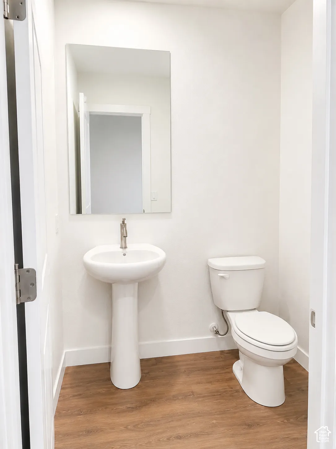White walls, wood-finish flooring, rectangular wall-mounted mirror, white pedestal sink with single-handle faucet, and white two-piece toilet