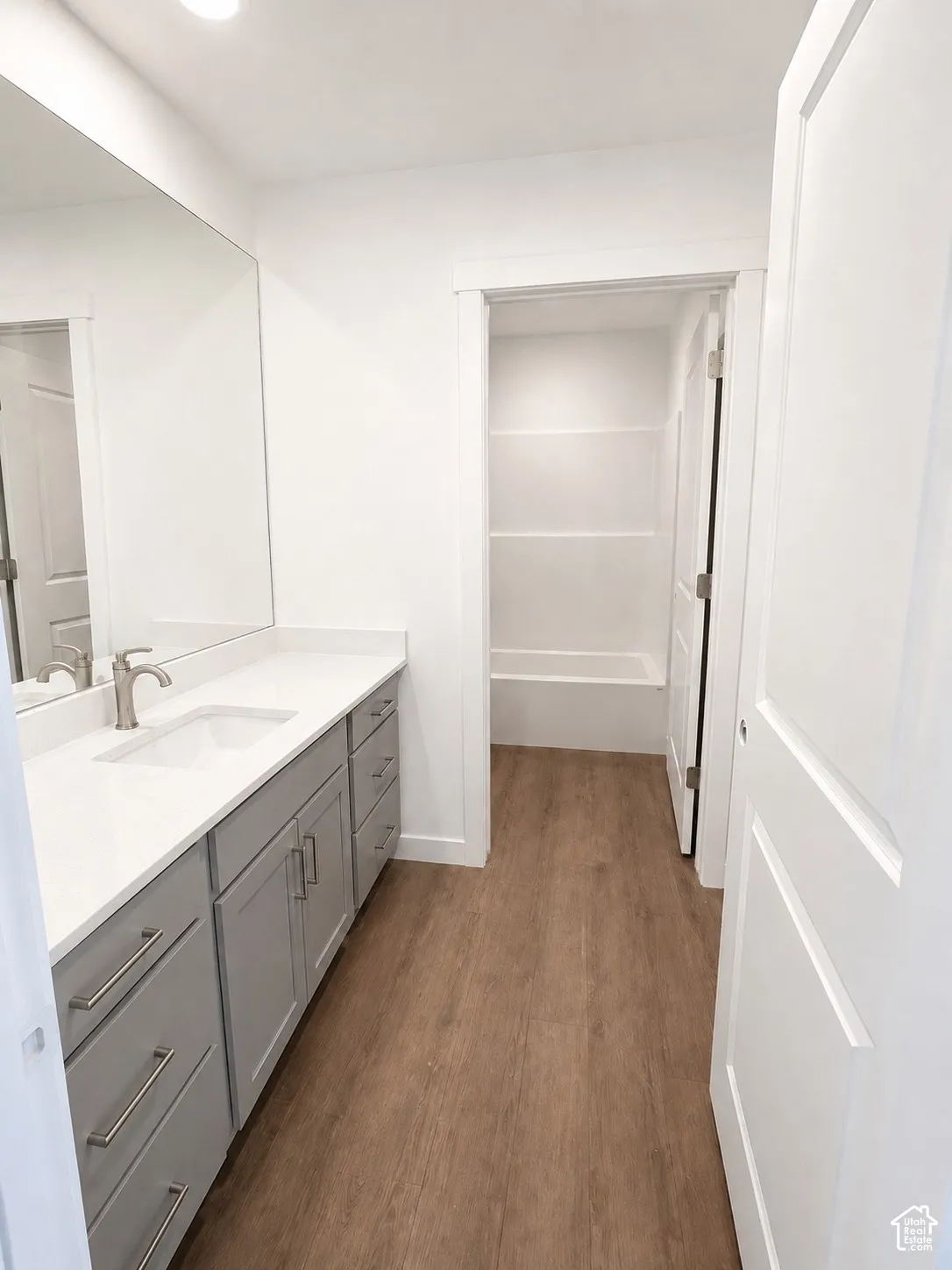 Wood-finish flooring, a white vanity countertop, gray cabinetry, and a built-in tub with shelving